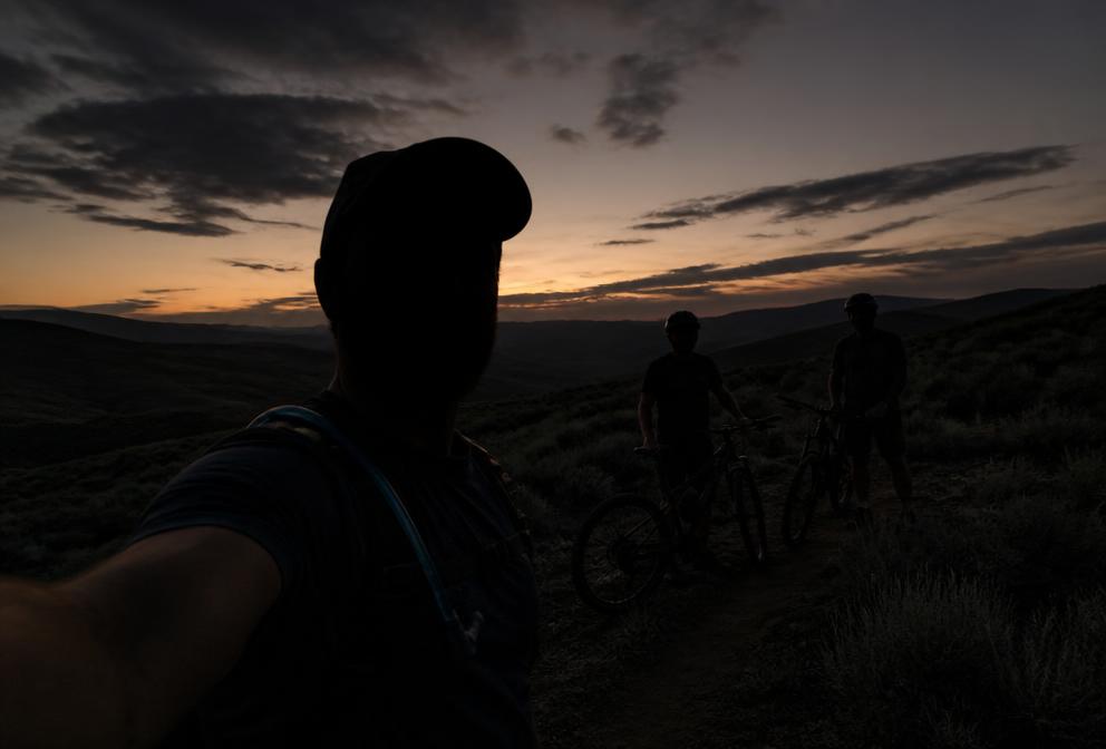 Silhouette of a rider taking a trail selfie with two others and mountain bikes at dusk