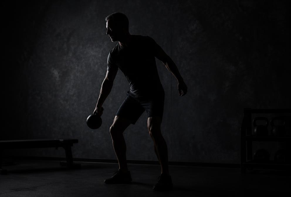 Silhouette of a lean athletic man training with a kettlebell in a dim gym