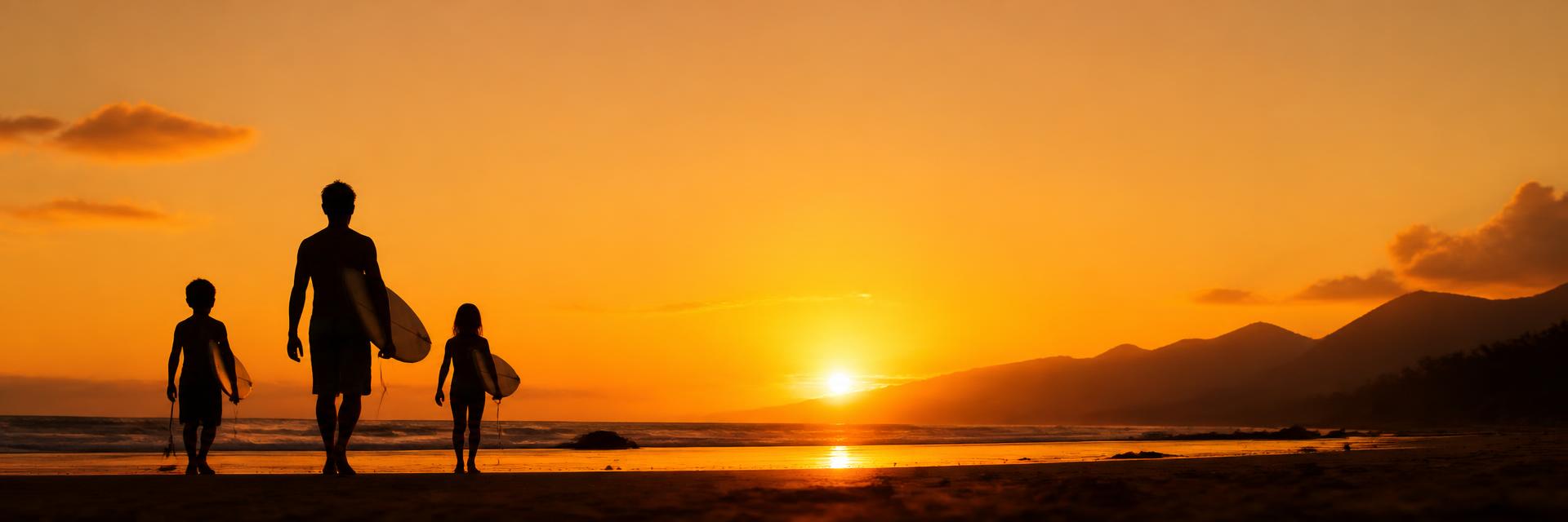 Silhouette of a dad and two kids carrying surfboards toward the ocean at sunset